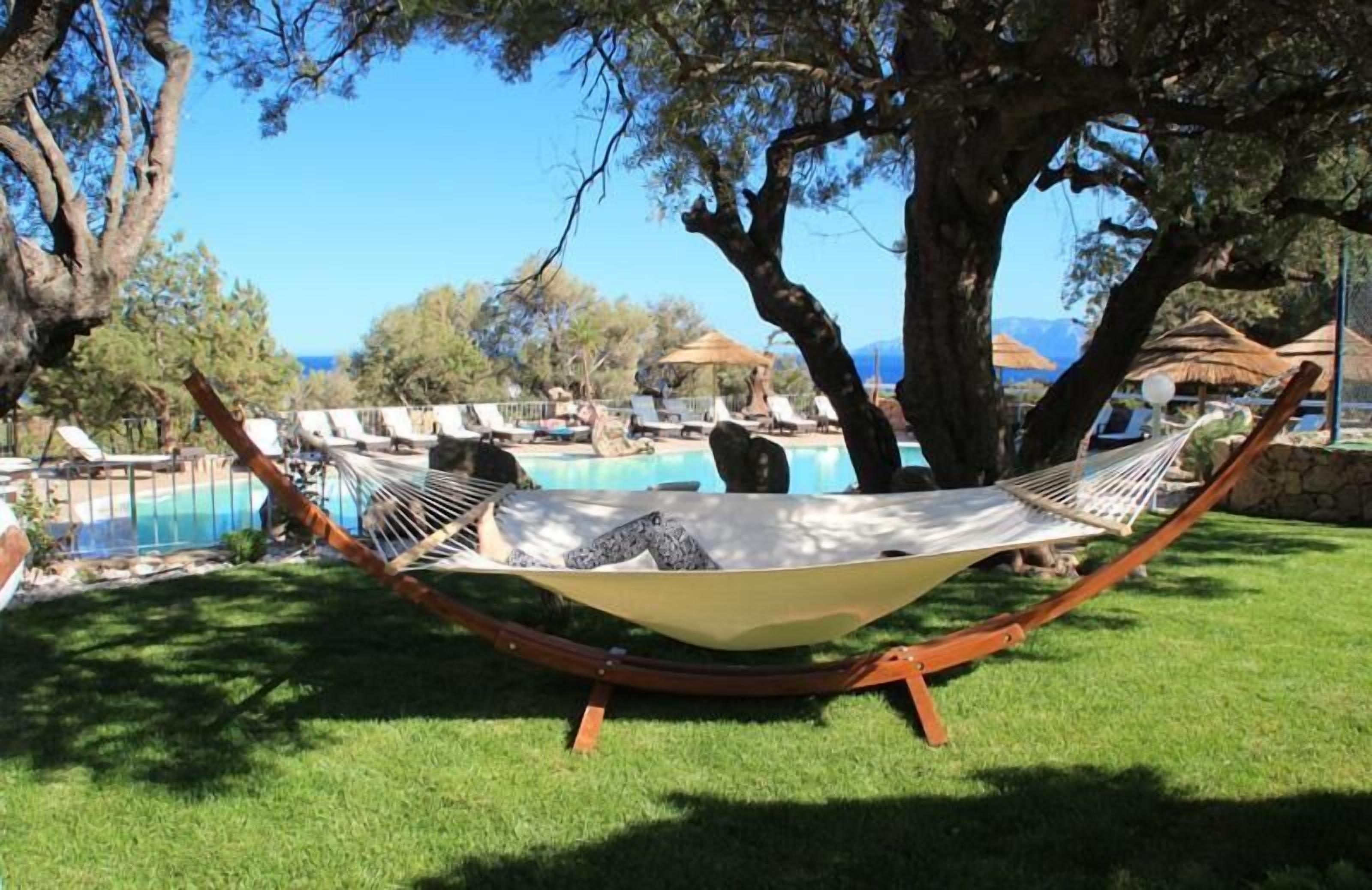 Una hamaca bajo un árbol, con una piscina y sillas de descanso al fondo.