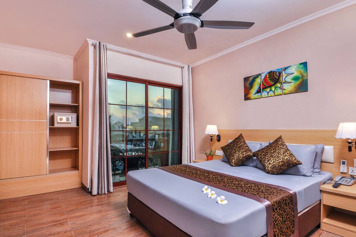 Quarto de hotel com cama, ventilador de teto, pintura, mesa de cabeceira e vista através de uma porta de vidro.
