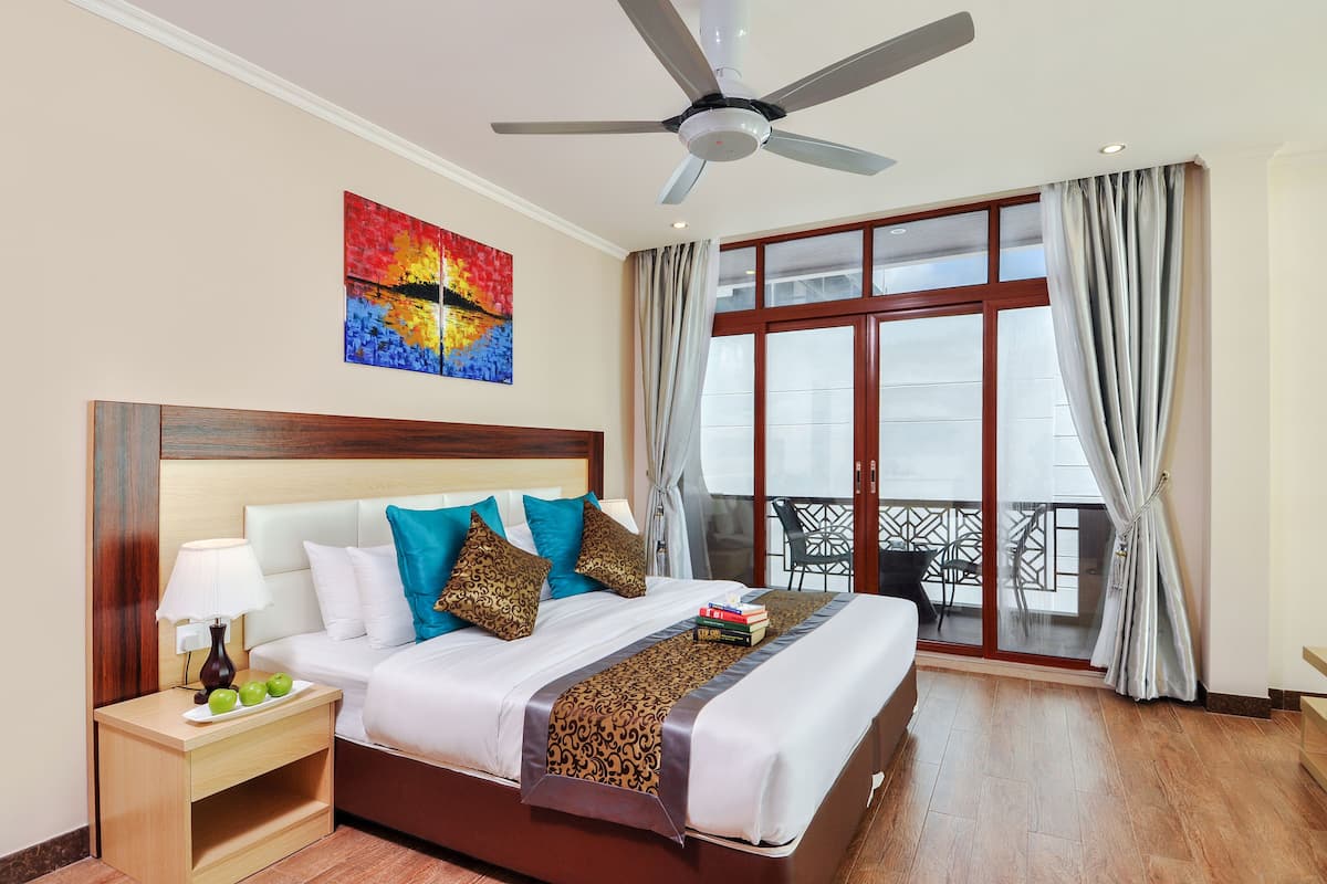 Quarto de hotel com cama, mesa de cabeceira com abajur e maçãs, ventoinha no teto, janela grande com cortinas e varanda com cadeiras.