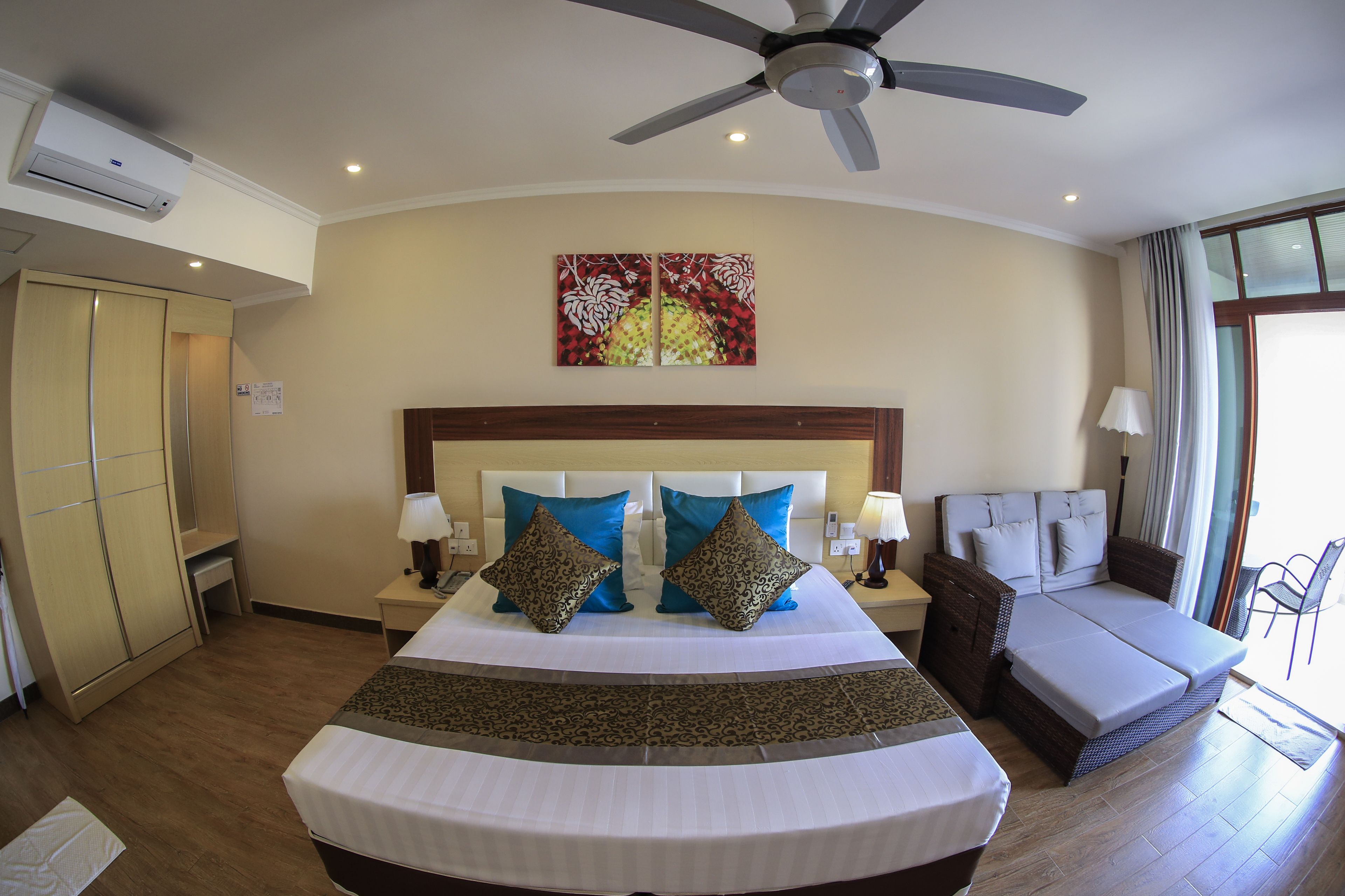 A hotel room with a bed, a ceiling fan, a window with curtains, a nightstand with a lamp, and a wall-mounted air conditioner.