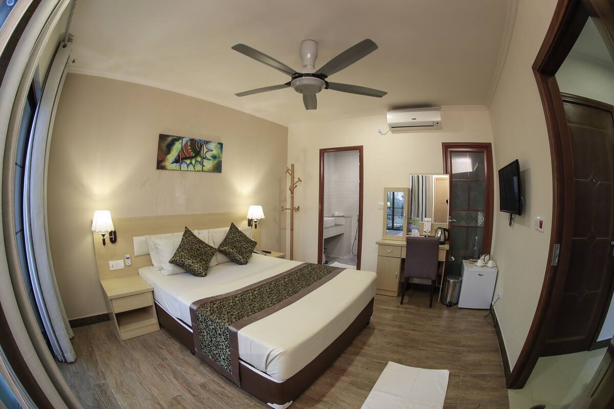 Quarto de hotel com cama, mesas de cabeceira, escrivaninha, cadeira, ventoinha no teto e aparelho de ar condicionado, além de uma televisão.