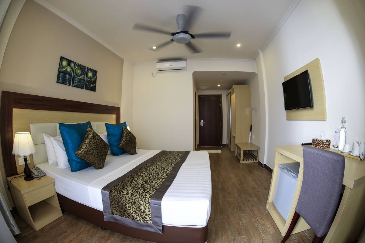 Quarto de hotel com cama, mesa de cabeceira, abajur, aparelho de ar condicionado, televisão, escrivaninha, cadeira e obra de arte na parede.