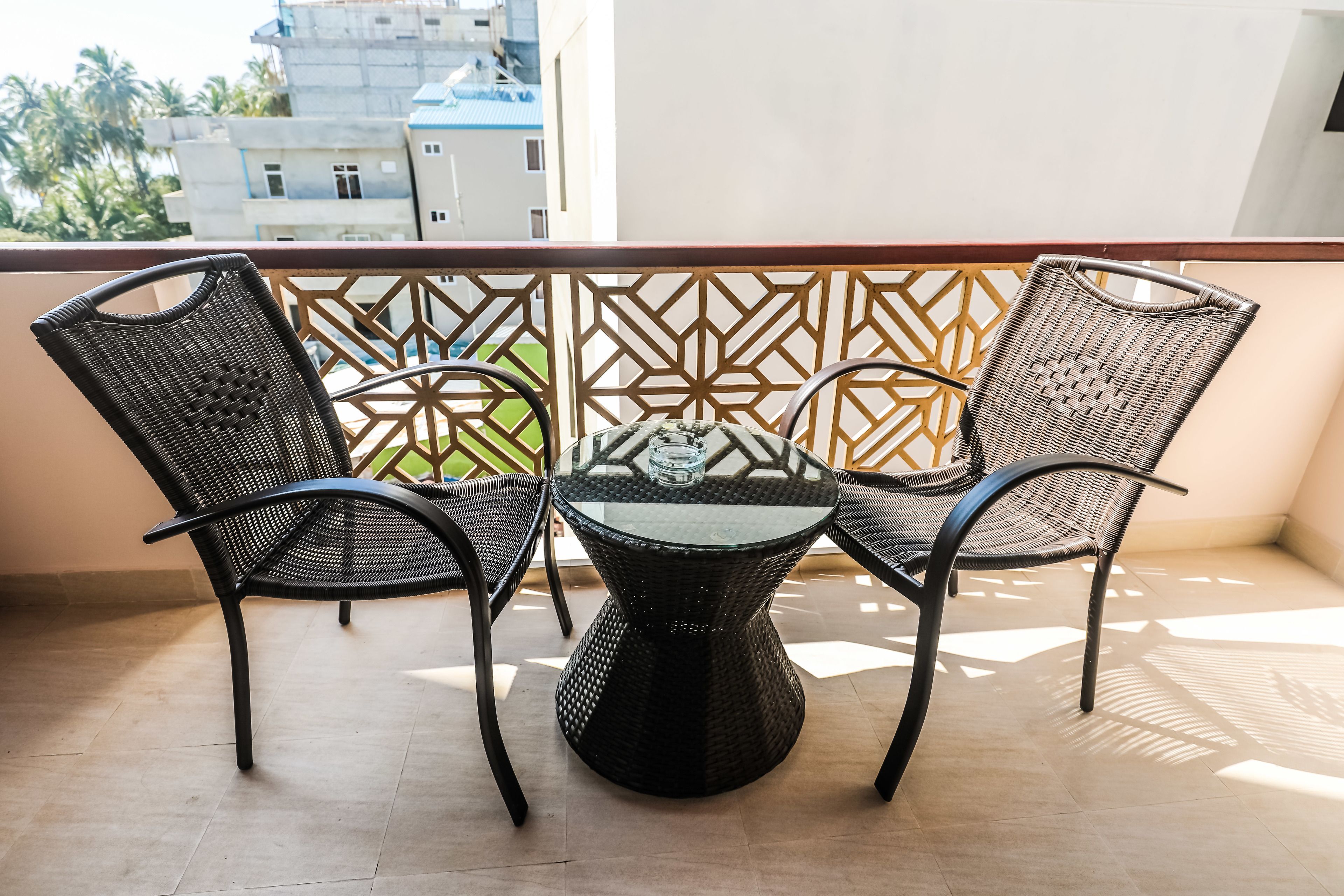 A balcony with a table and four chairs, a geometric patterned railing, and a view of buildings and palm trees in the background.