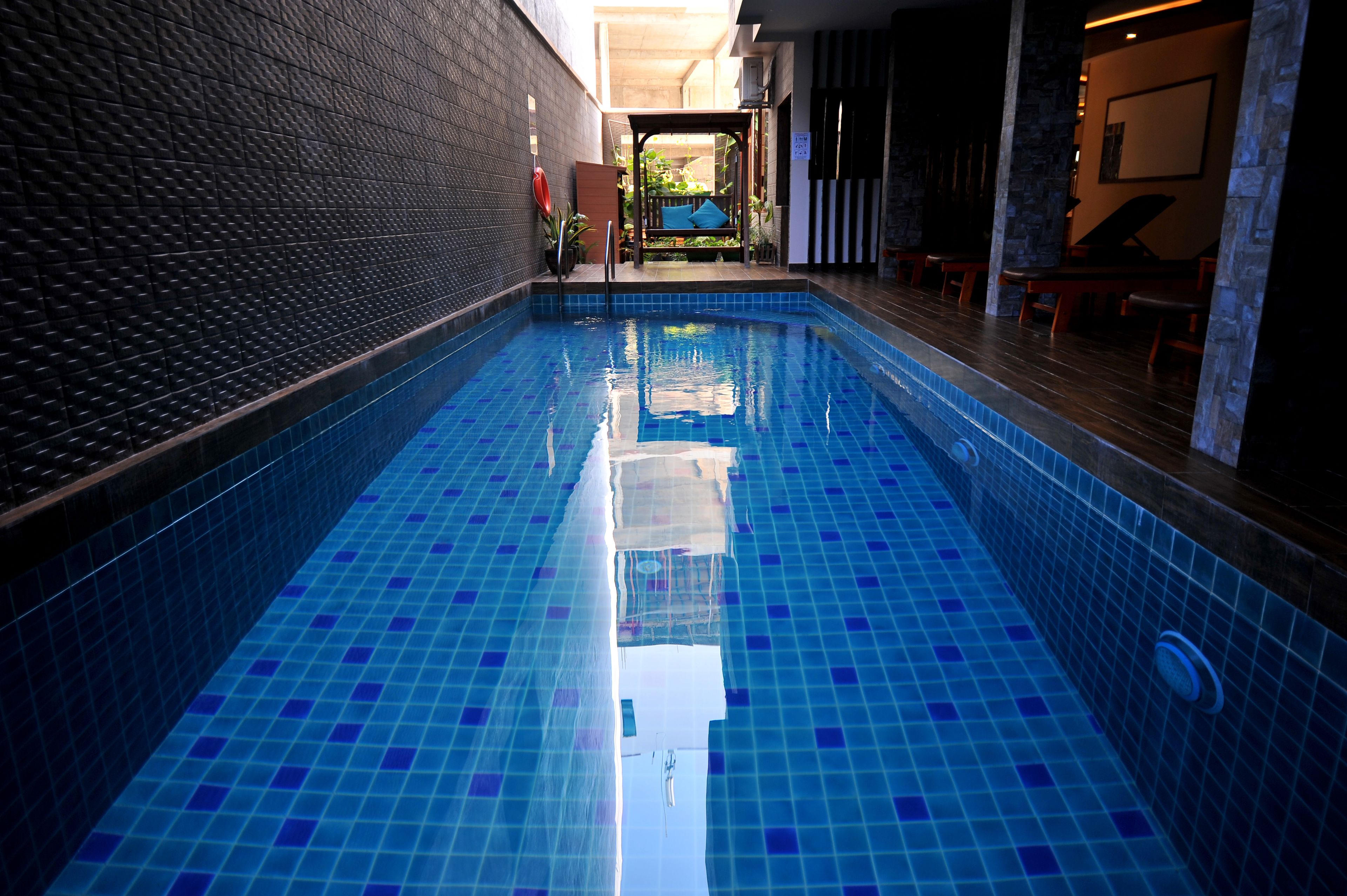 A swimming pool with blue tiles, a wooden deck, and a covered seating area with plants.