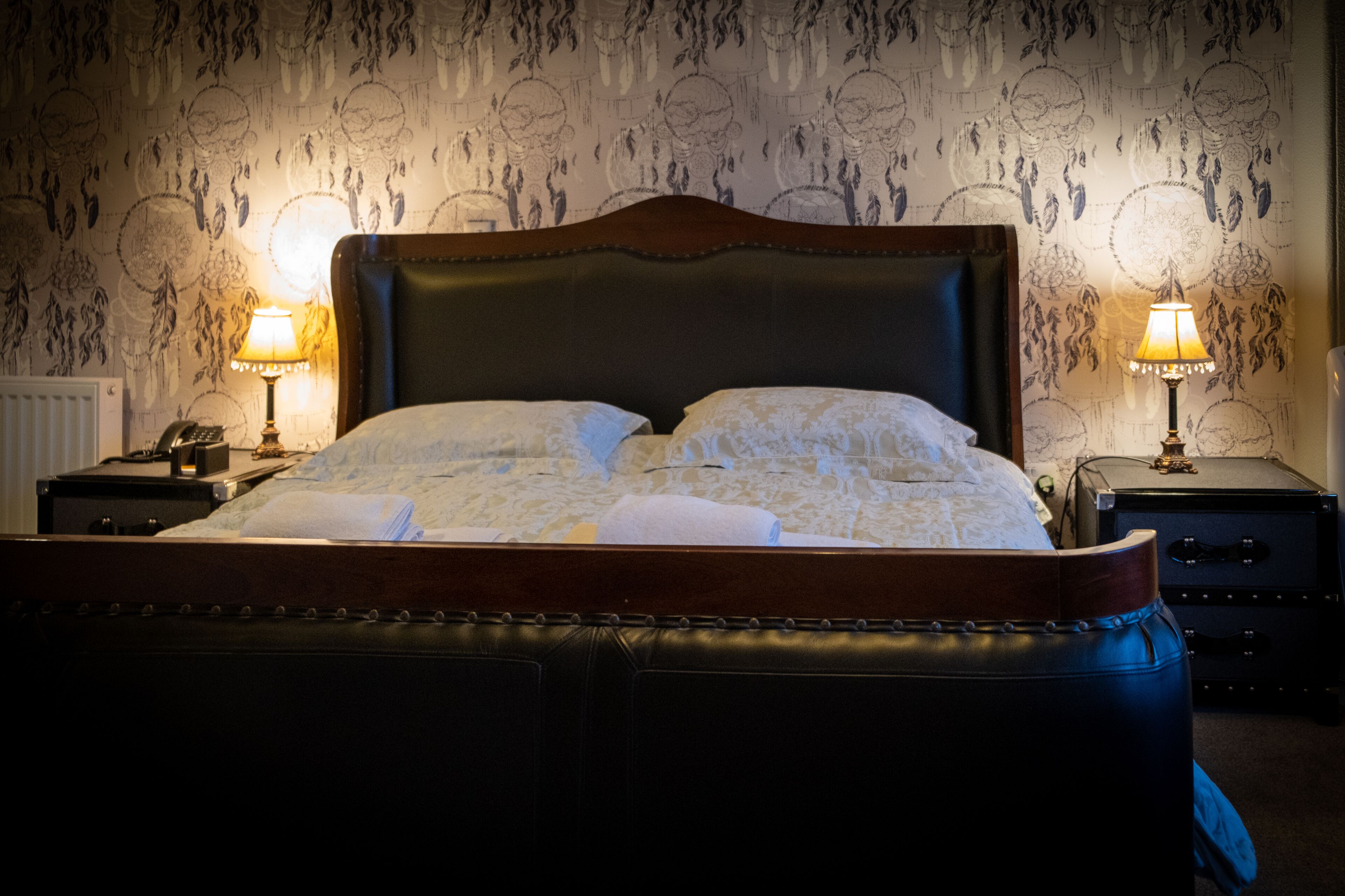 A neatly made bed with a wooden headboard, two bedside lamps, and a patterned wallpaper.