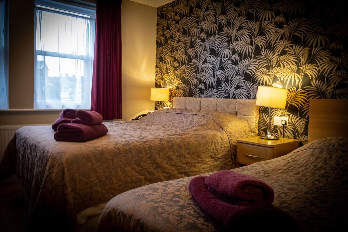 Een hotelkamer met twee bedden, een raam met gordijnen, behang met een patroon en twee bedlampen.