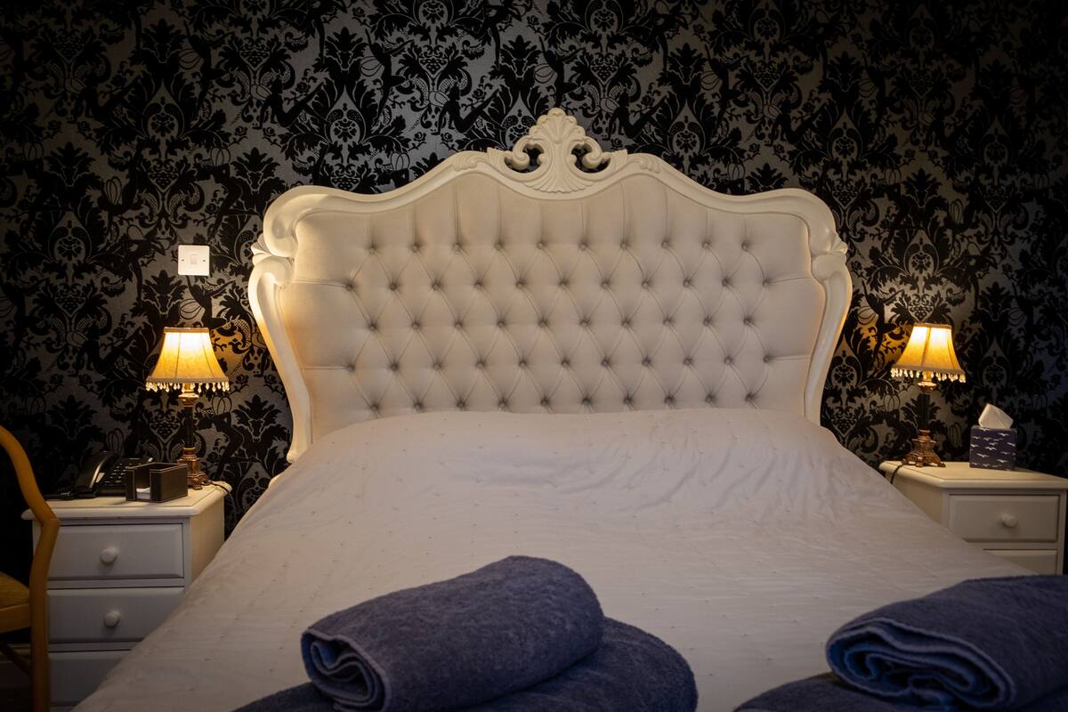 Een netjes opgemaakt bed met een gestoffeerd hoofdbord, twee bedlampjes en opgerolde handdoeken op het bed.