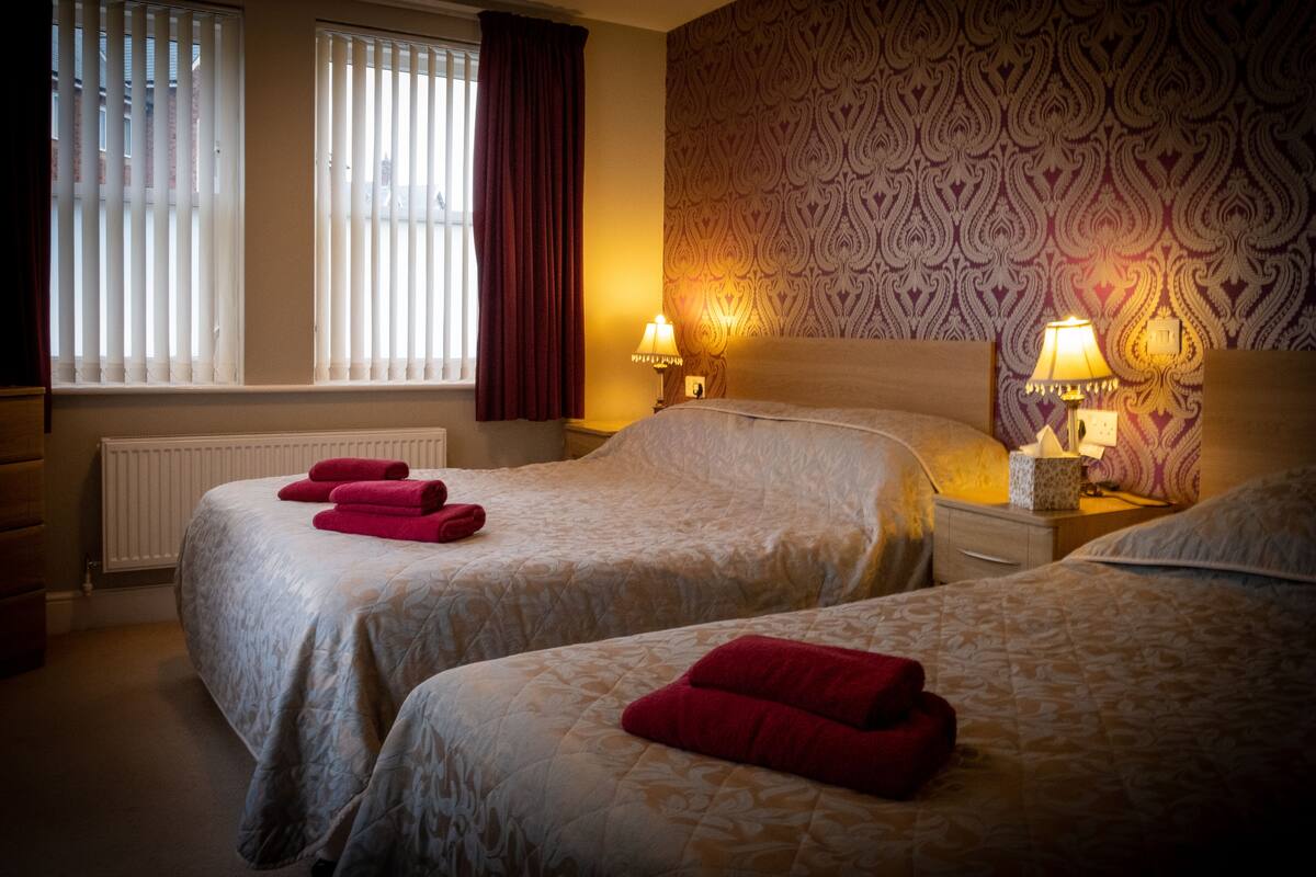 Een hotelkamer met twee bedden, een radiator, een raam met zonwering en behang met een patroon.