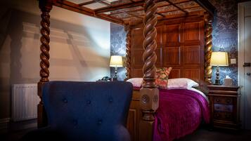A traditional four-poster bed with a wooden headboard, a blue upholstered chair, and a wooden nightstand with a lamp.