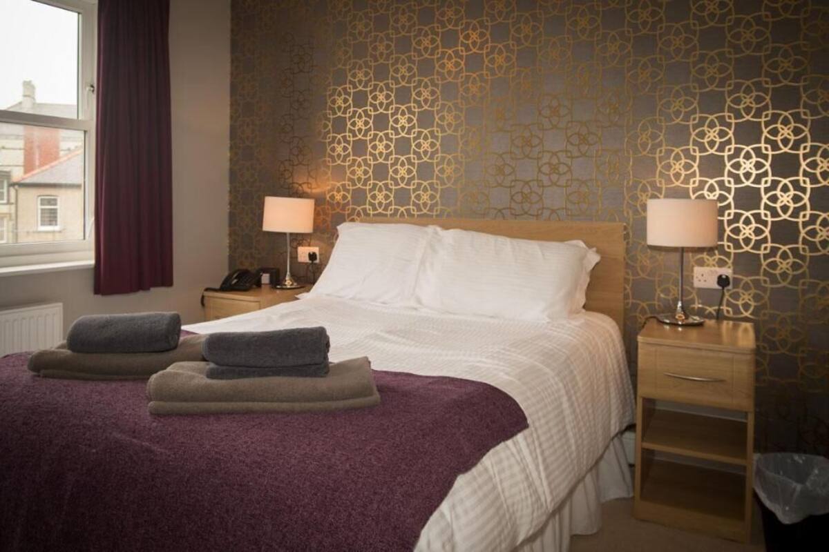 Een hotelkamer met een bed, twee bedlampen, een nachtkastje, een raam met gordijnen en behang met een patroon.