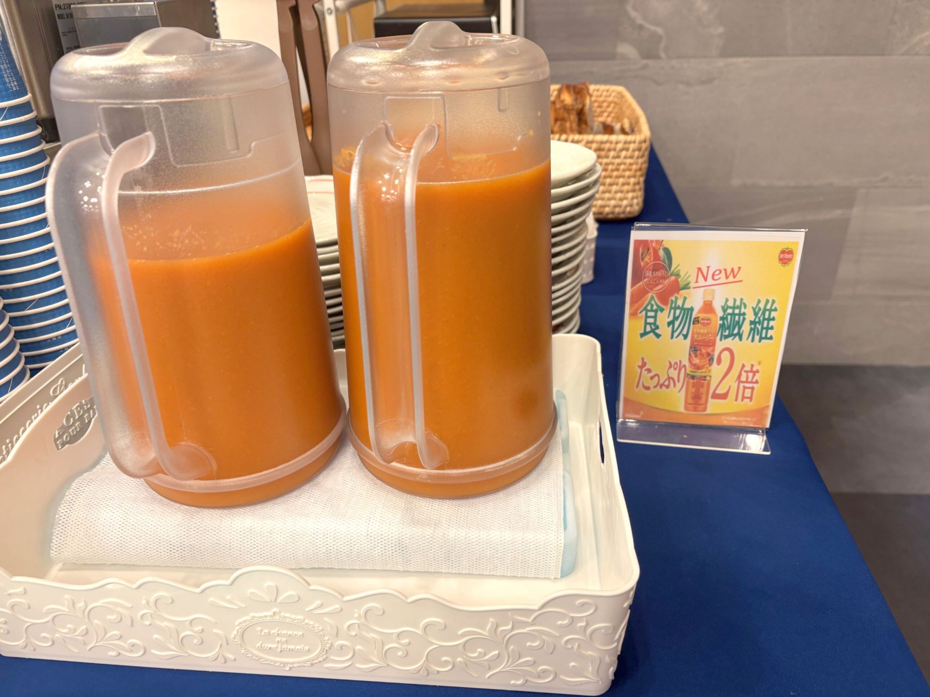 Dos vasos de plástico transparente llenos de jugo de naranja sobre una bandeja blanca con un borde decorativo. Un letrero con texto japonés se ve al fondo.