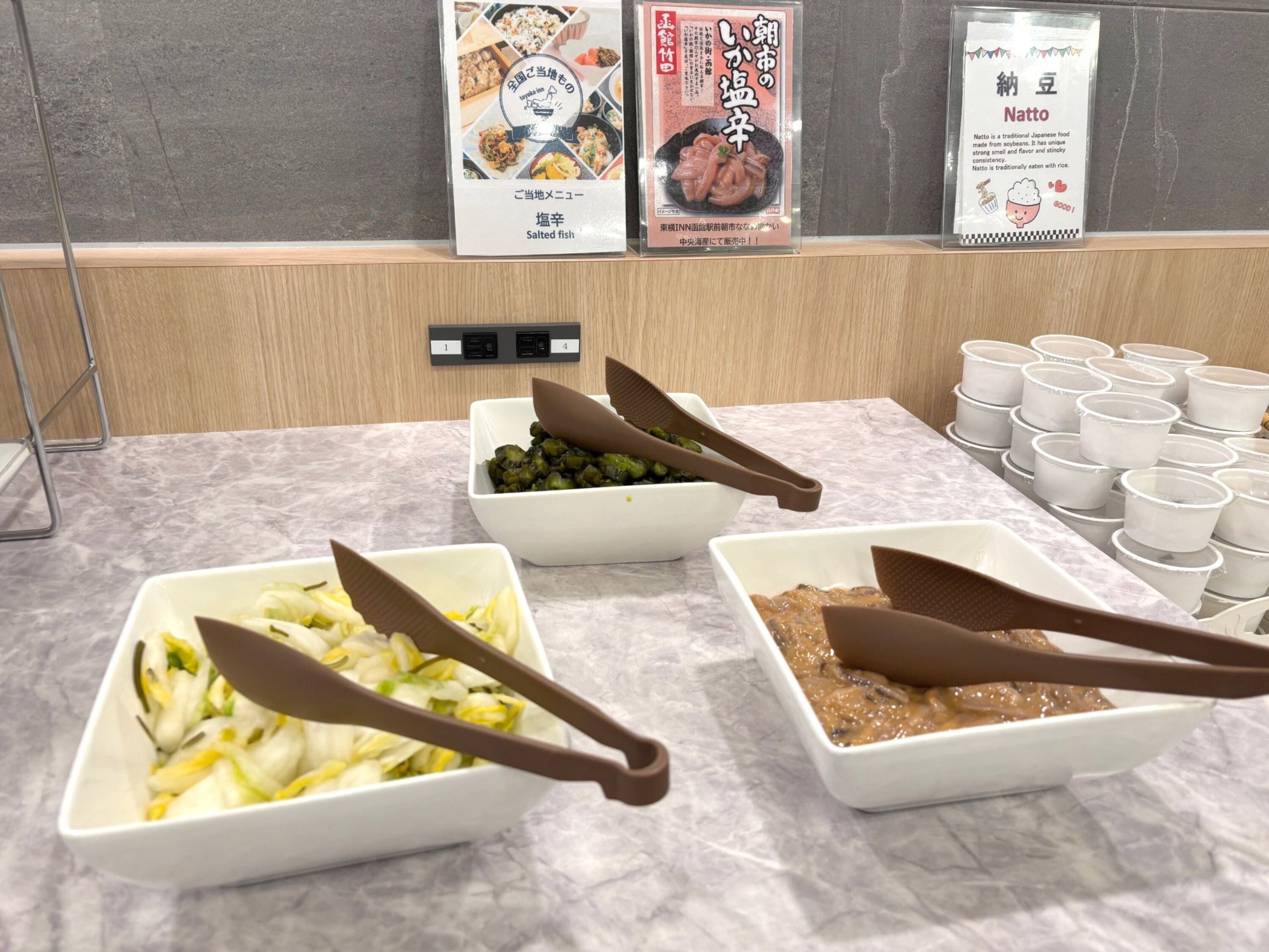 Una mesa de buffet con diversos platos, incluyendo un tazón de verduras cortadas, un tazón de vegetales verdes y un tazón de salsa marrón. También hay varios vasos de plástico y un letrero con texto japonés.