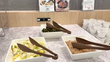 Una mesa de buffet con diversos platos, incluyendo un tazón de verduras cortadas, un tazón de vegetales verdes y un tazón de salsa marrón. También hay varios vasos de plástico y un letrero con texto japonés.
