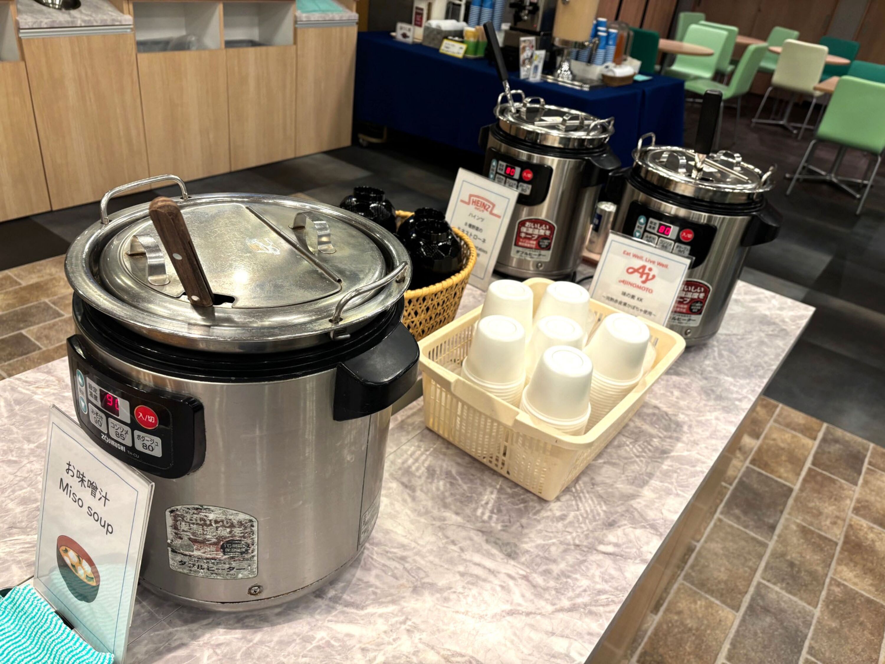 A kitchen area with a large electric pressure cooker, a basket of disposable cups, and a sign indicating a menu item.