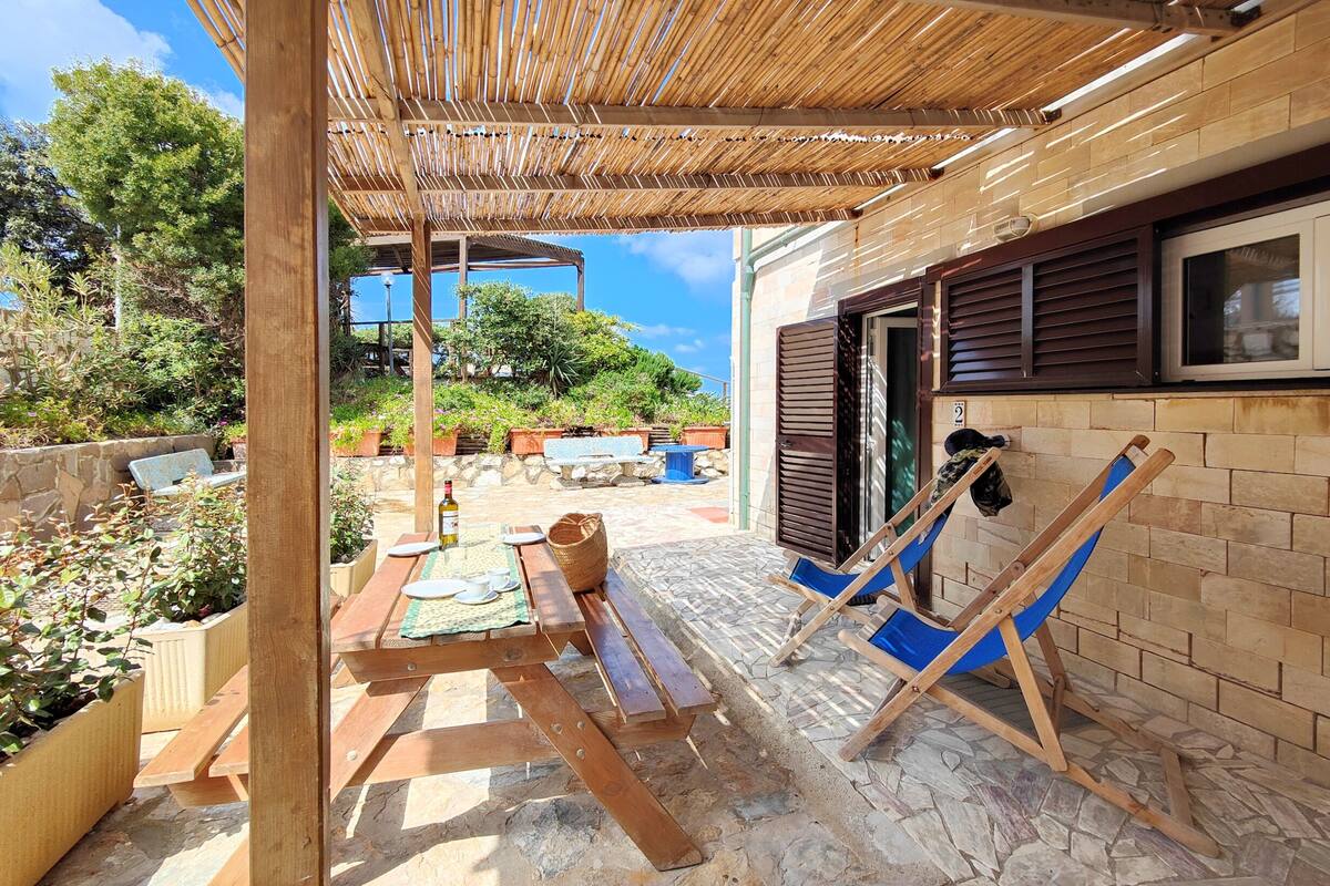 A covered outdoor patio with wooden furniture, a stone floor, and a view of a building with a window and shutters.