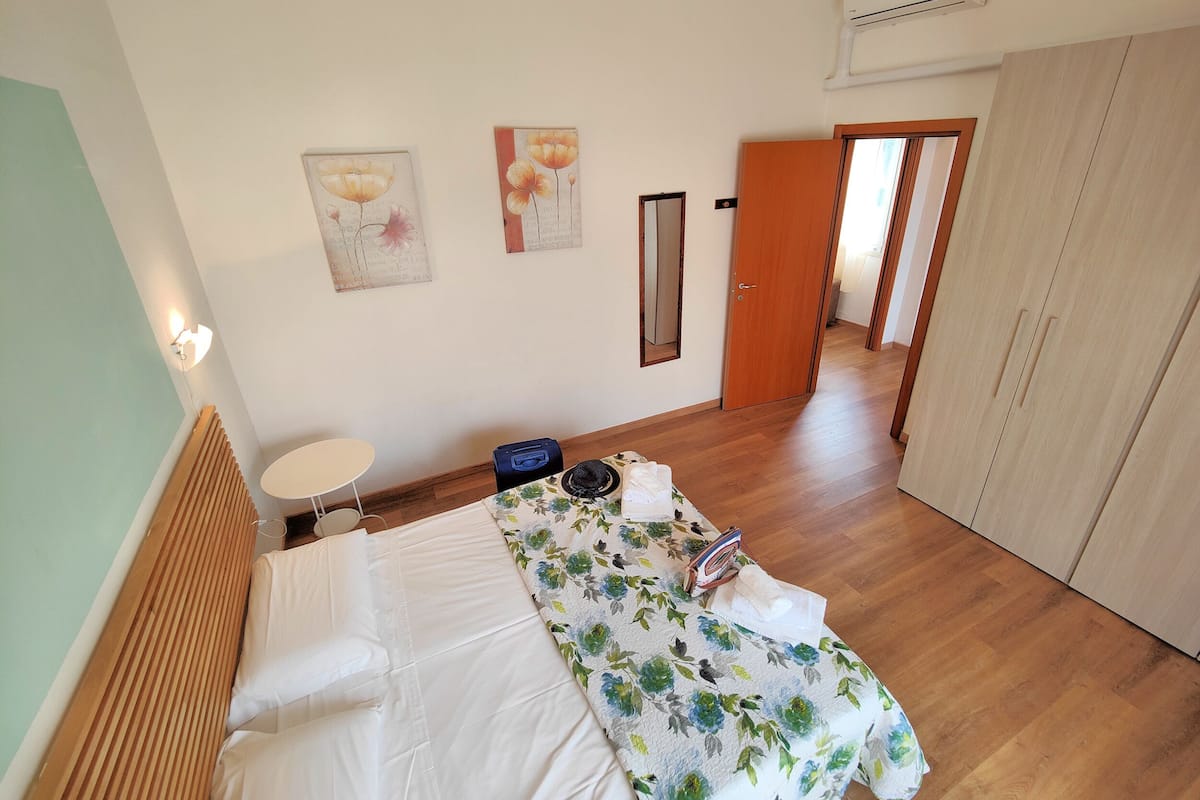 A bedroom with a bed, a small table, a suitcase, and a door leading to another room.