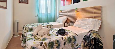A bedroom with two beds, a window with sheer curtains, and a wall with four framed artworks.