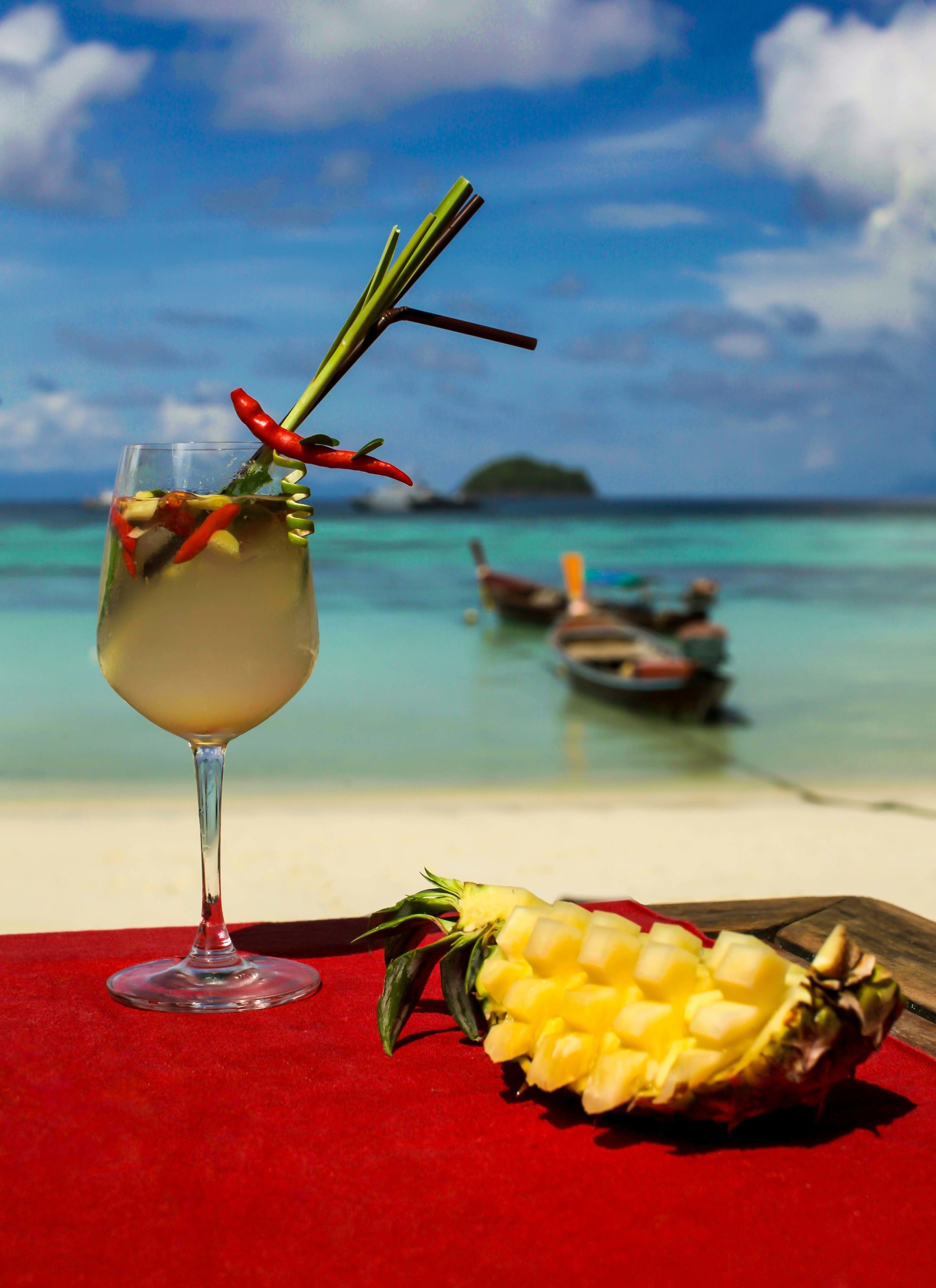 Ein Getränk mit Trinkhalm und Ananas auf einer roten Tischdecke am Strand, im Hintergrund sind Boote zu sehen.