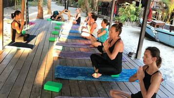 Un grupo de personas sentadas sobre esterillas de yoga bajo una estructura de madera, posiblemente un pabellón, con una playa y palmeras al fondo.