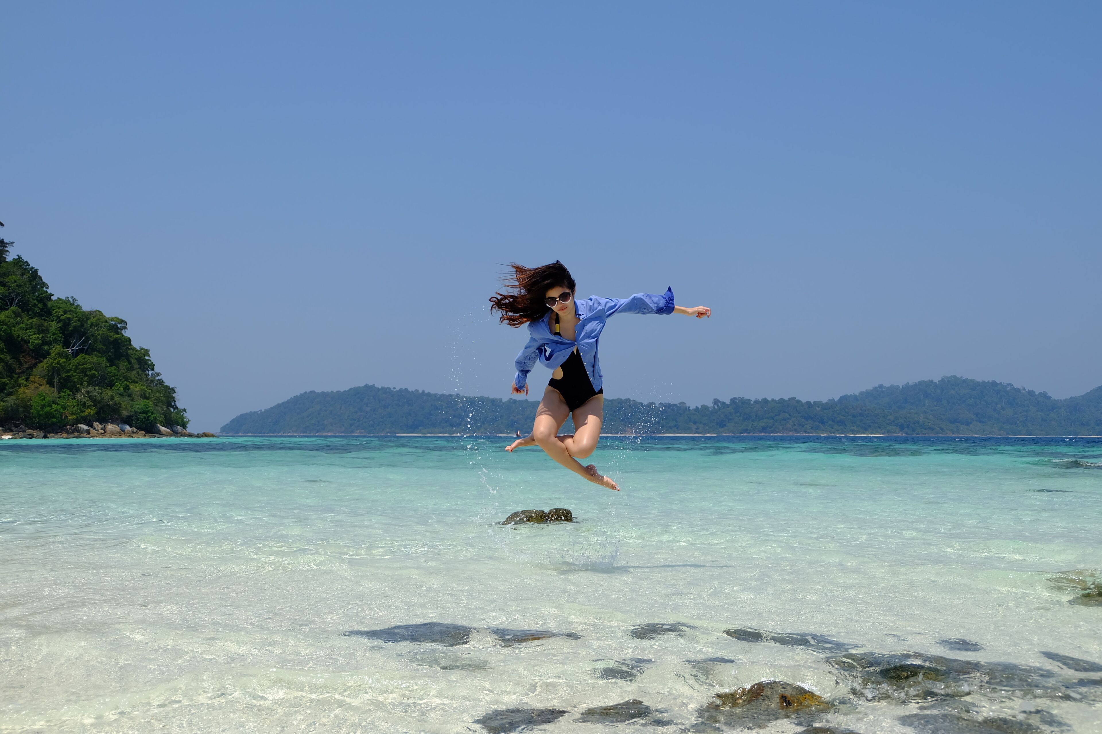A person jumping into clear turquoise water with a rocky shore and green hills in the background.