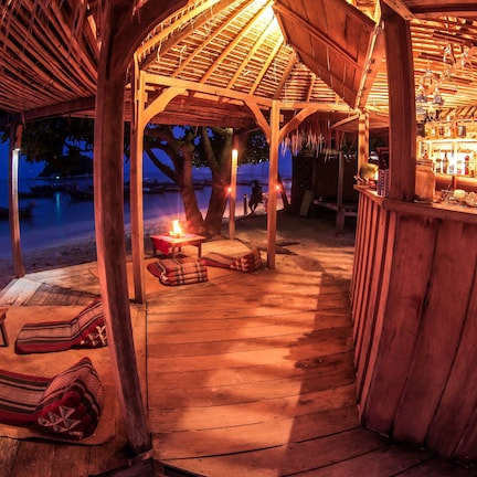 A wooden beach bar with a thatched roof, lounge chairs, and a bar area with bottles.