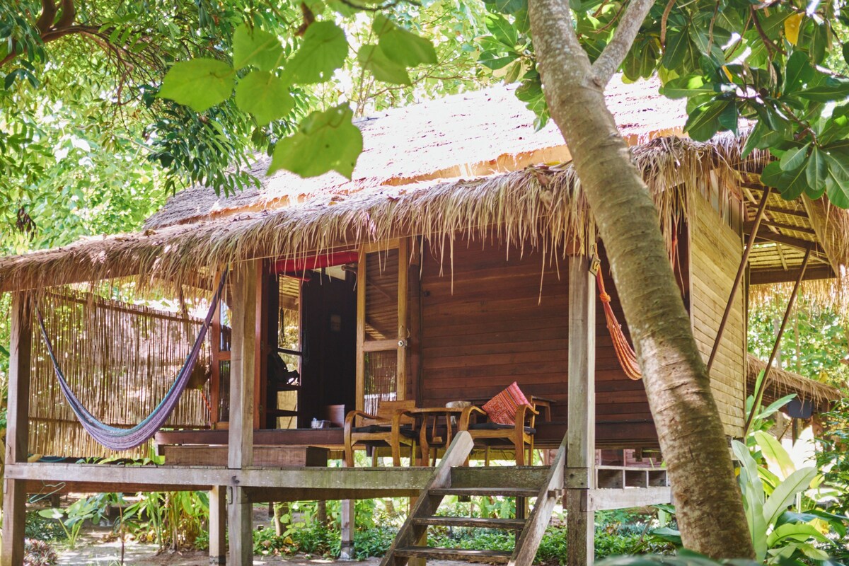 A wooden cabin with a thatched roof, a hammock, and a wooden deck.