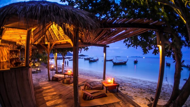 A wooden beach hut with a thatched roof, furnished with cushions and a table, overlooking a calm sea with boats. - Castaway Resort Koh Lipe (Koh Lipe)