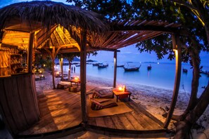 A wooden beach hut with a thatched roof, furnished with cushions and a table, overlooking a calm sea with boats. - Castaway Resort Koh Lipe (Koh Lipe)