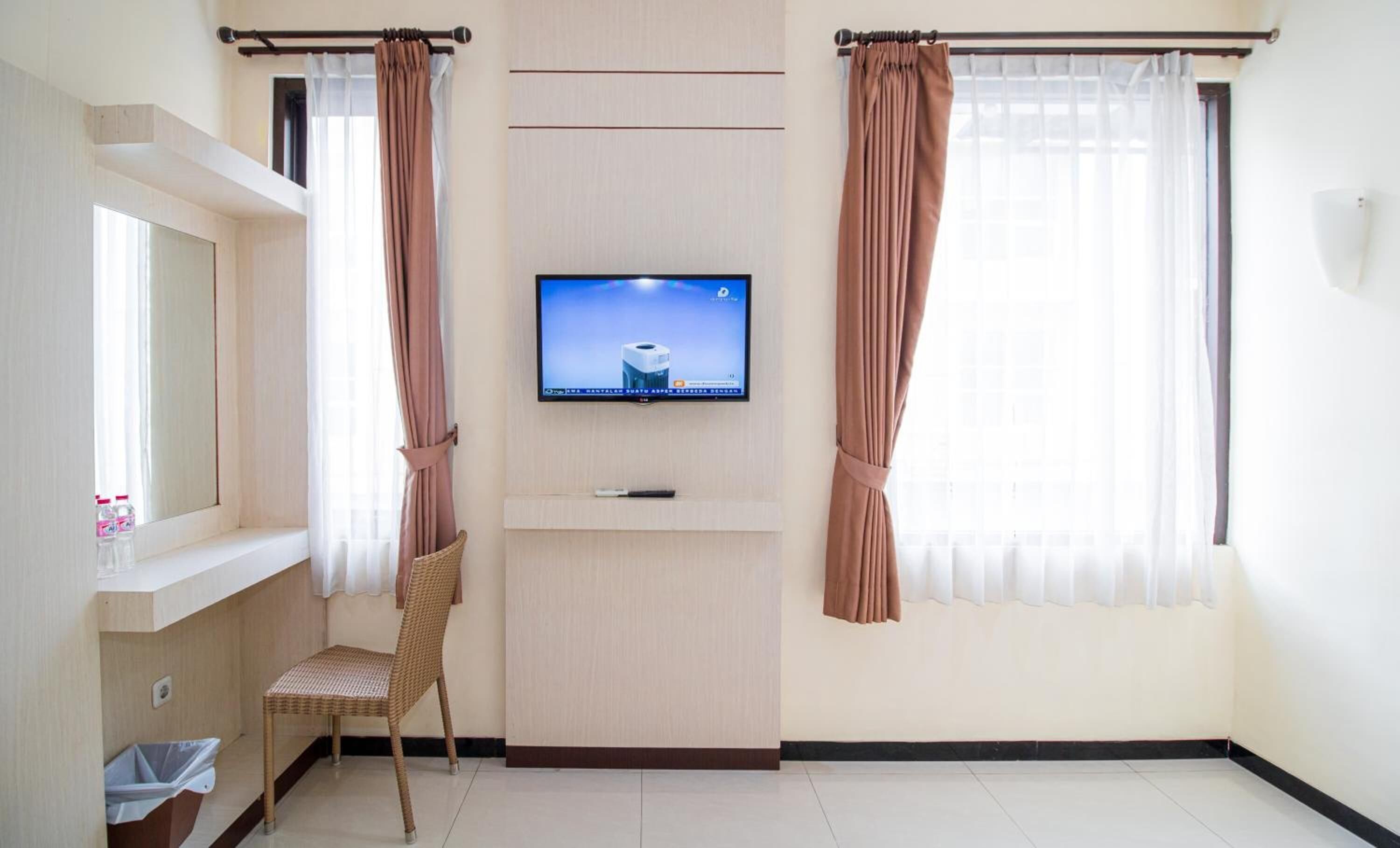 A hotel room with a flat-screen TV mounted on the wall, a wicker chair, a small trash bin, and a window with curtains.