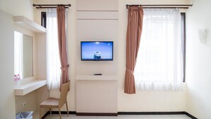 A hotel room with a flat-screen TV mounted on the wall, a wicker chair, a small trash bin, and a window with curtains. - Lily Guest House (Malang)