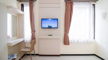 A hotel room with a flat-screen TV mounted on the wall, a wicker chair, a small trash bin, and a window with curtains.