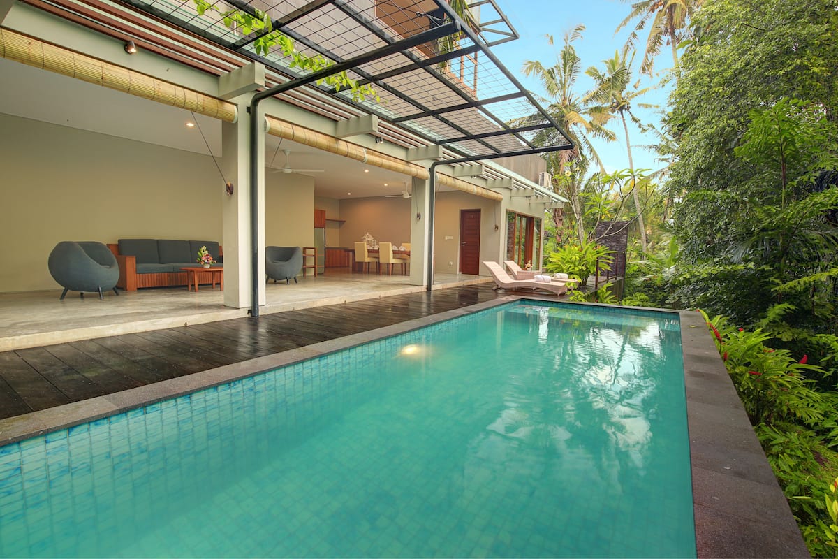A modern indoor swimming pool with a glass-enclosed patio area, a dining area, and lush greenery surrounding the space.