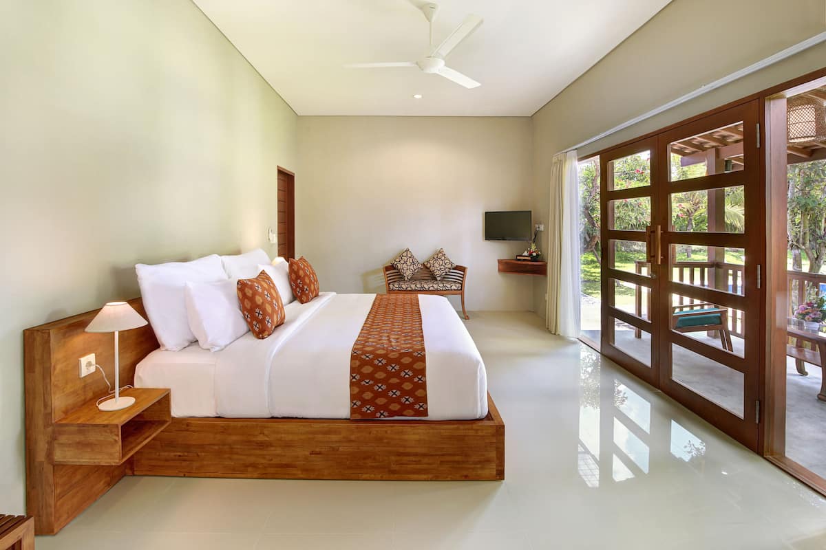 A spacious bedroom with a large bed, wooden headboard, a bedside table with a lamp, and a TV on the wall.
