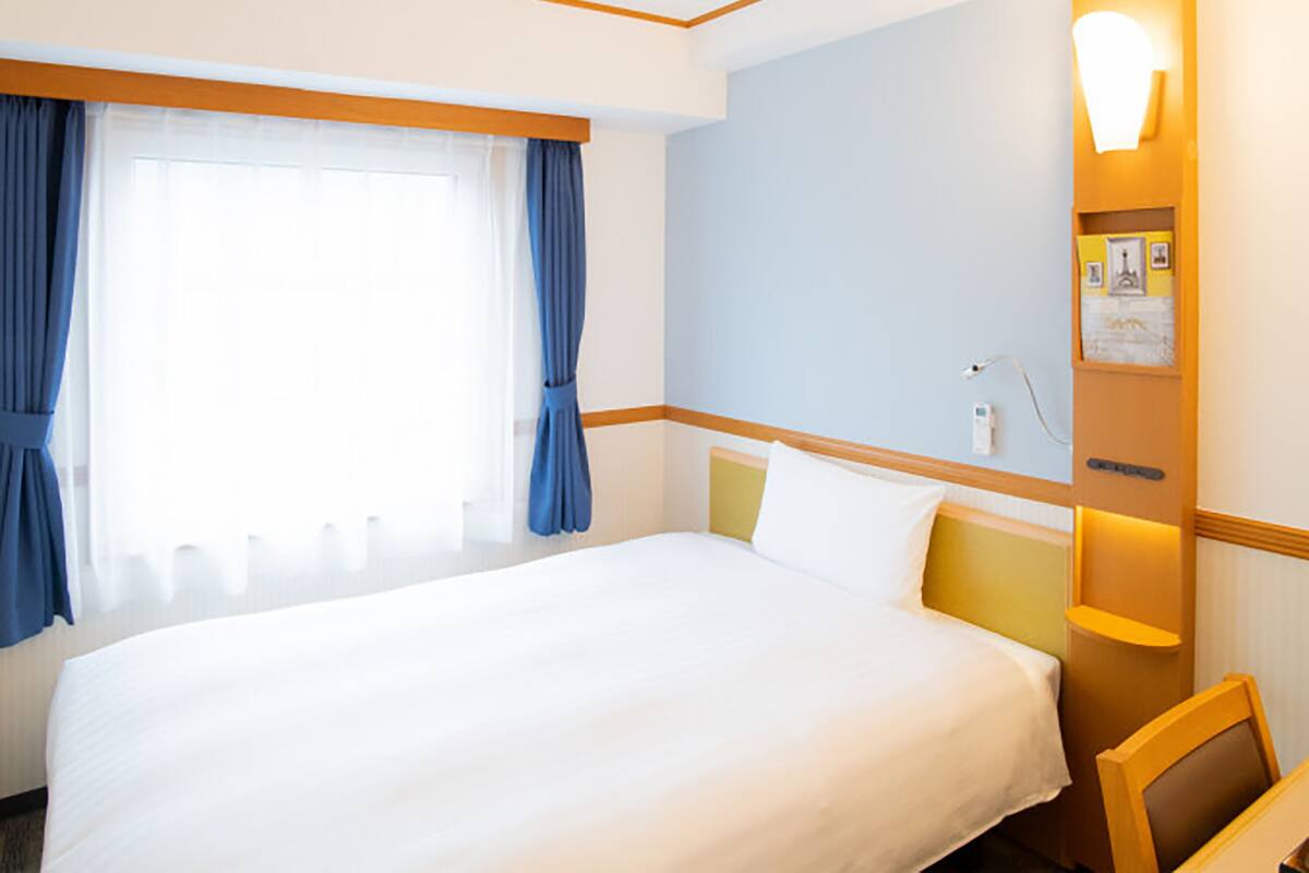 Una habitación de hotel con una cama grande, una mesita de noche, una silla, un ventanal con cortinas y una lámpara fijada a la pared.