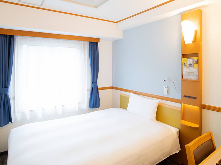 Una habitación de hotel con una cama grande, una mesita de noche, una silla, un ventanal con cortinas y una lámpara fijada a la pared.