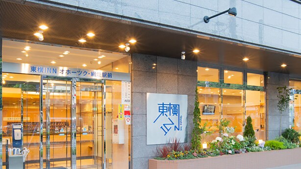 Entrada de hotel con puertas de vidrio, un cartel con caracteres japoneses y una jardinera de ladrillo con plantas.