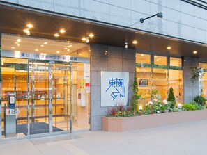Entrada de hotel con puertas de vidrio, un cartel con caracteres japoneses y una jardinera de ladrillo con plantas.