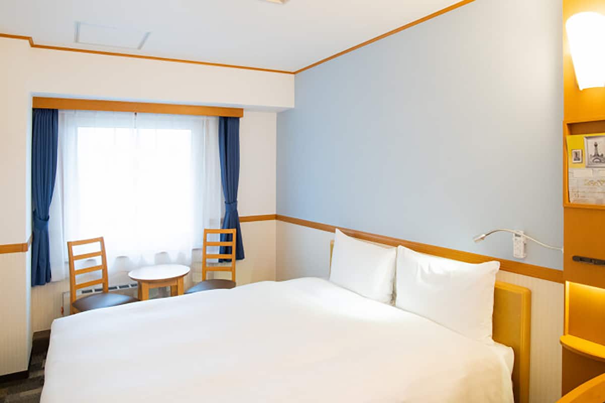 Habitación de hotel con una cama grande, una mesita pequeña con dos sillas, una ventana con cortinas y una lámpara fijada a la pared.