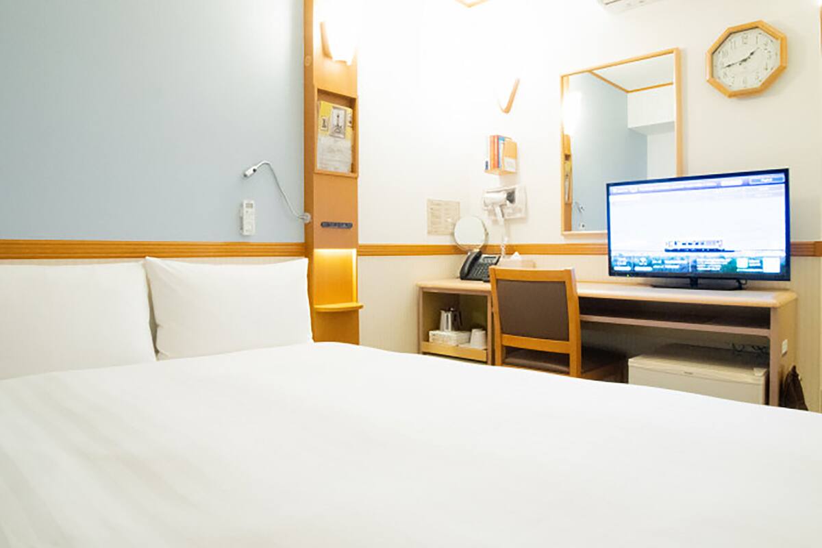 Una habitación de hotel con una cama, un escritorio con computadora, una silla, una lámpara de escritorio, un reloj y una estantería con libros.