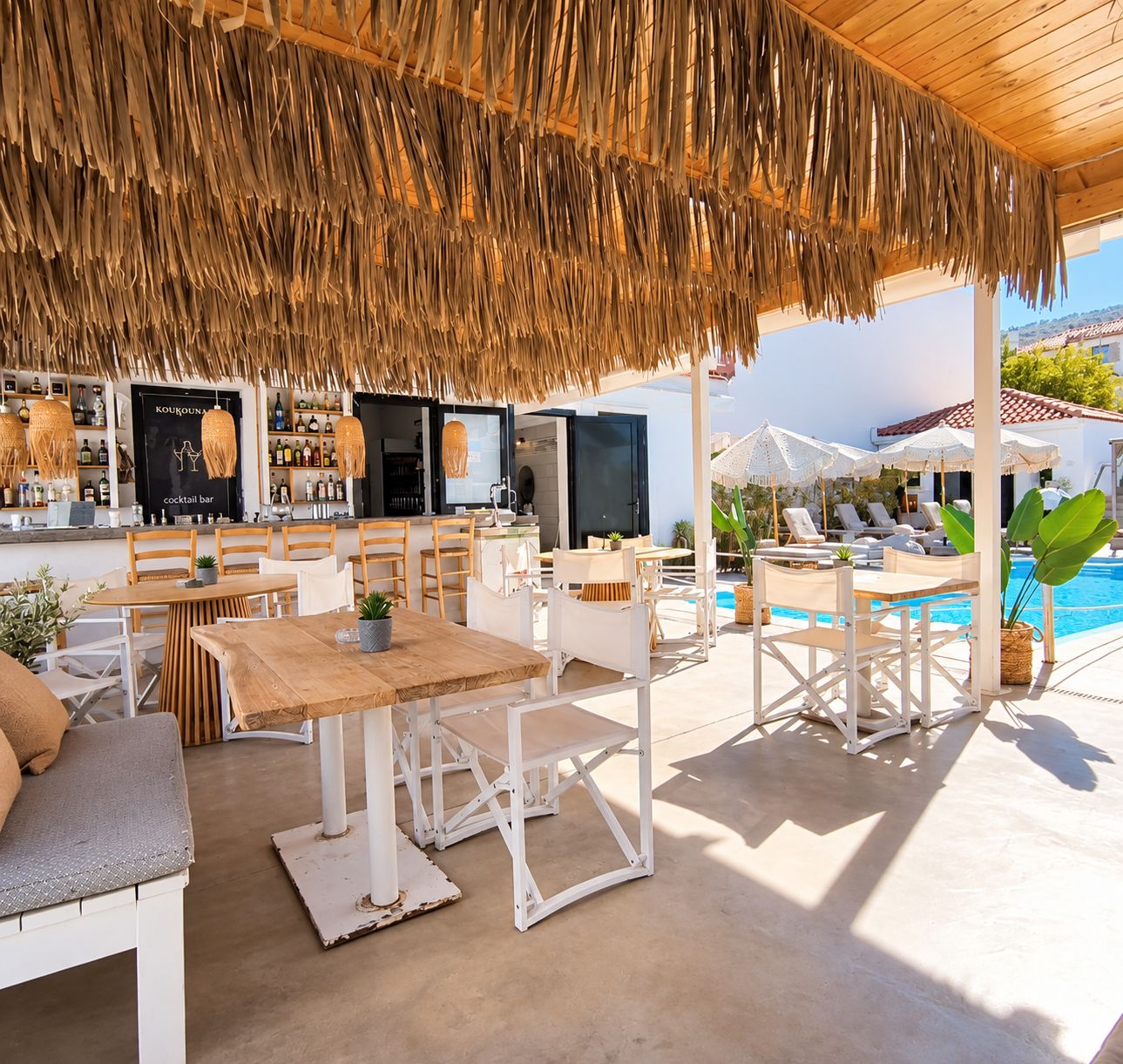 An outdoor seating area with tables and chairs, a bar with bottles, and a pool in the background.