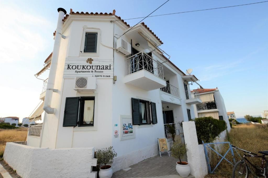 A two-story building with a balcony, a sign for 'Koukounari Apartments & Services,' and a contact number.