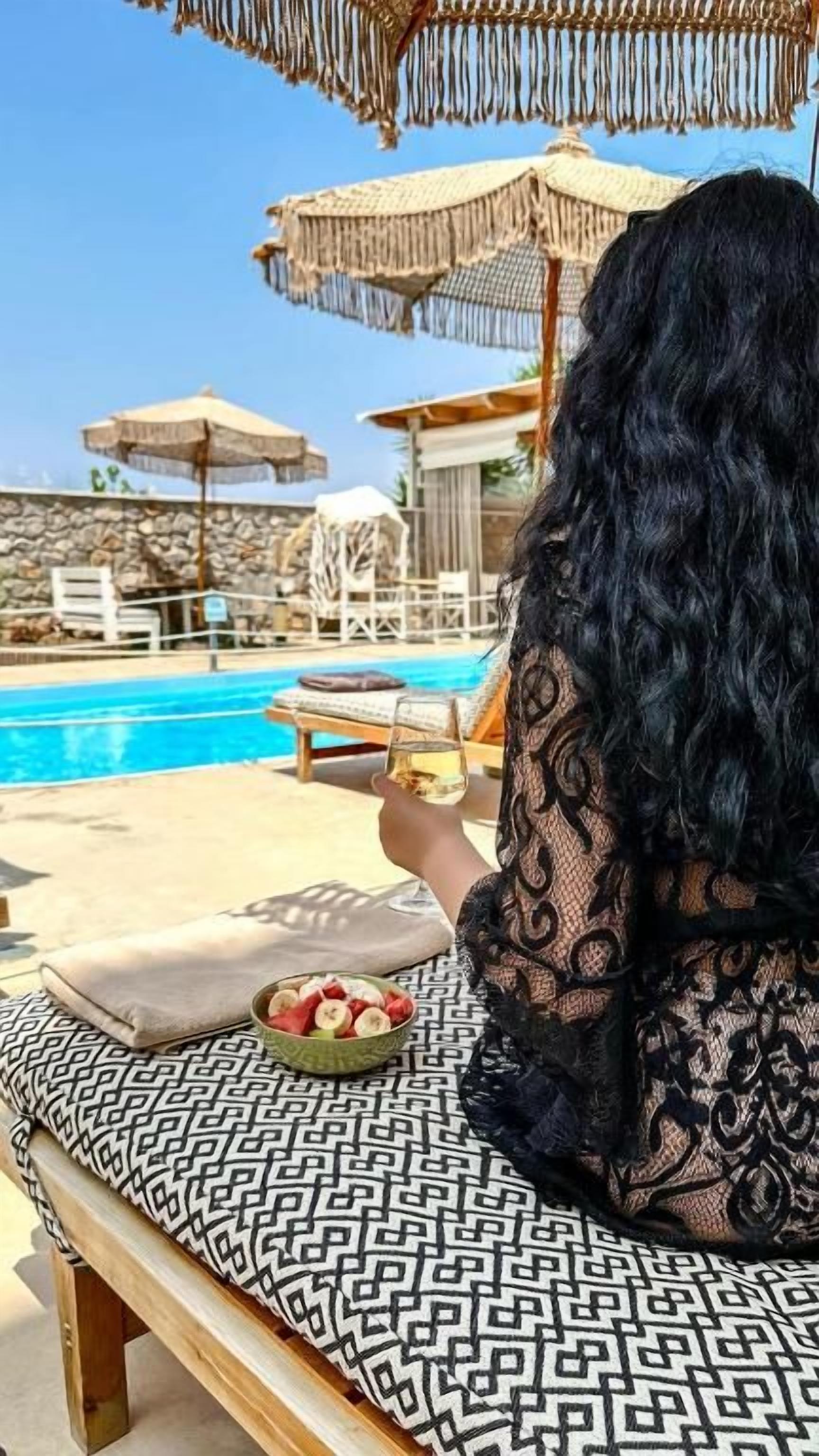 Una persona sentada en un banco estampado junto a una piscina, con un cuenco de fruta y una bebida.