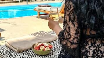 Una persona sentada en un banco estampado junto a una piscina, con un cuenco de fruta y una bebida.