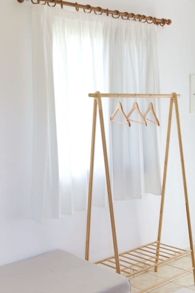 Una habitación con un tendedero de madera apoyado en una pared, una cama con colchón blanco y cortinas de tela ligera blanca.