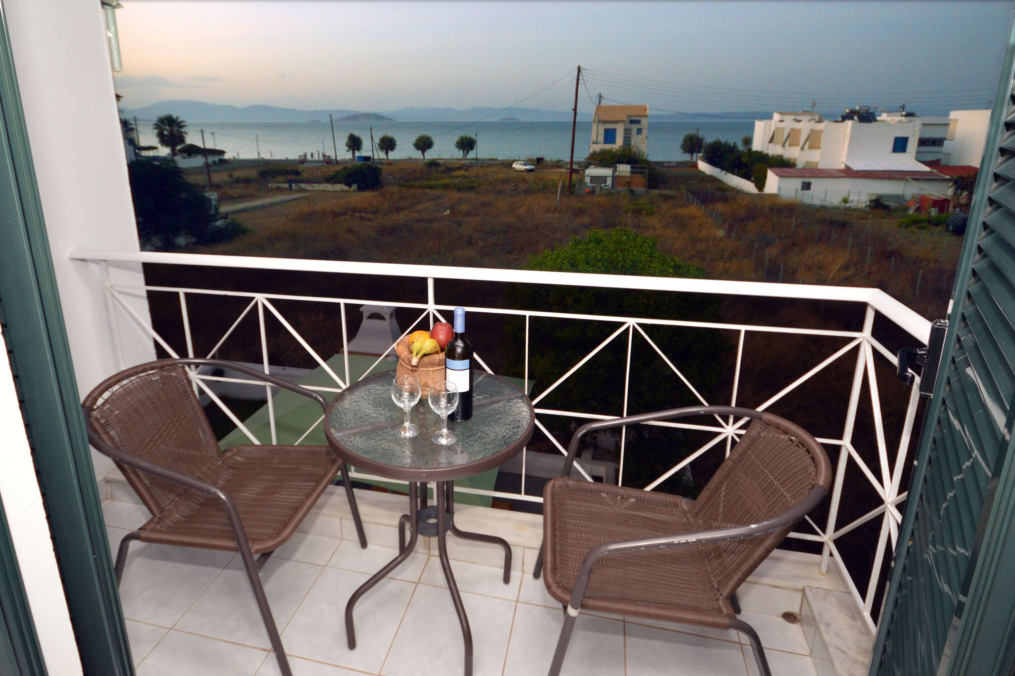A balcony with two wicker chairs and a small table, overlooking a coastal landscape with buildings and a view of the sea.