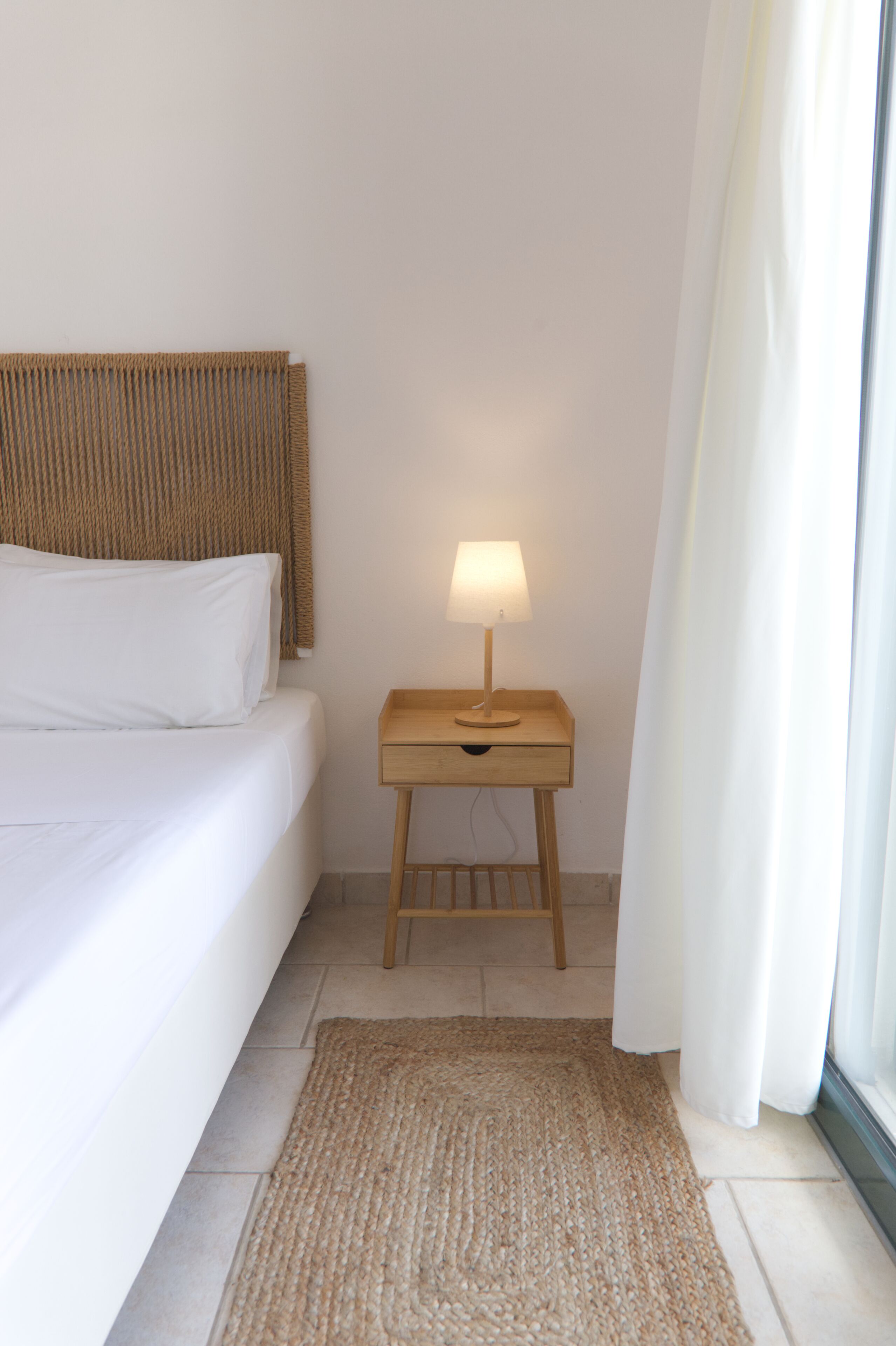 A neatly made bed with a wooden headboard, a bedside table with a lamp, and a woven rug on a tiled floor.