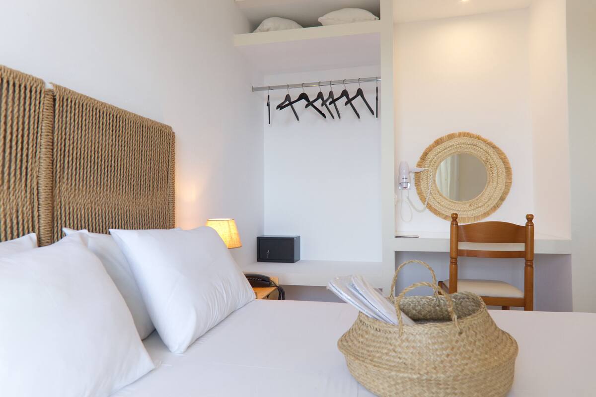 A neatly arranged hotel room with a bed, a bedside lamp, a wall-mounted mirror, a wooden chair, and a woven basket on the bed.