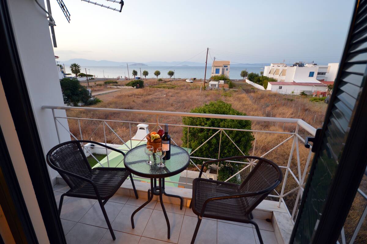 A balcony with two chairs and a table, overlooking a coastal area with buildings and a view of the sea.