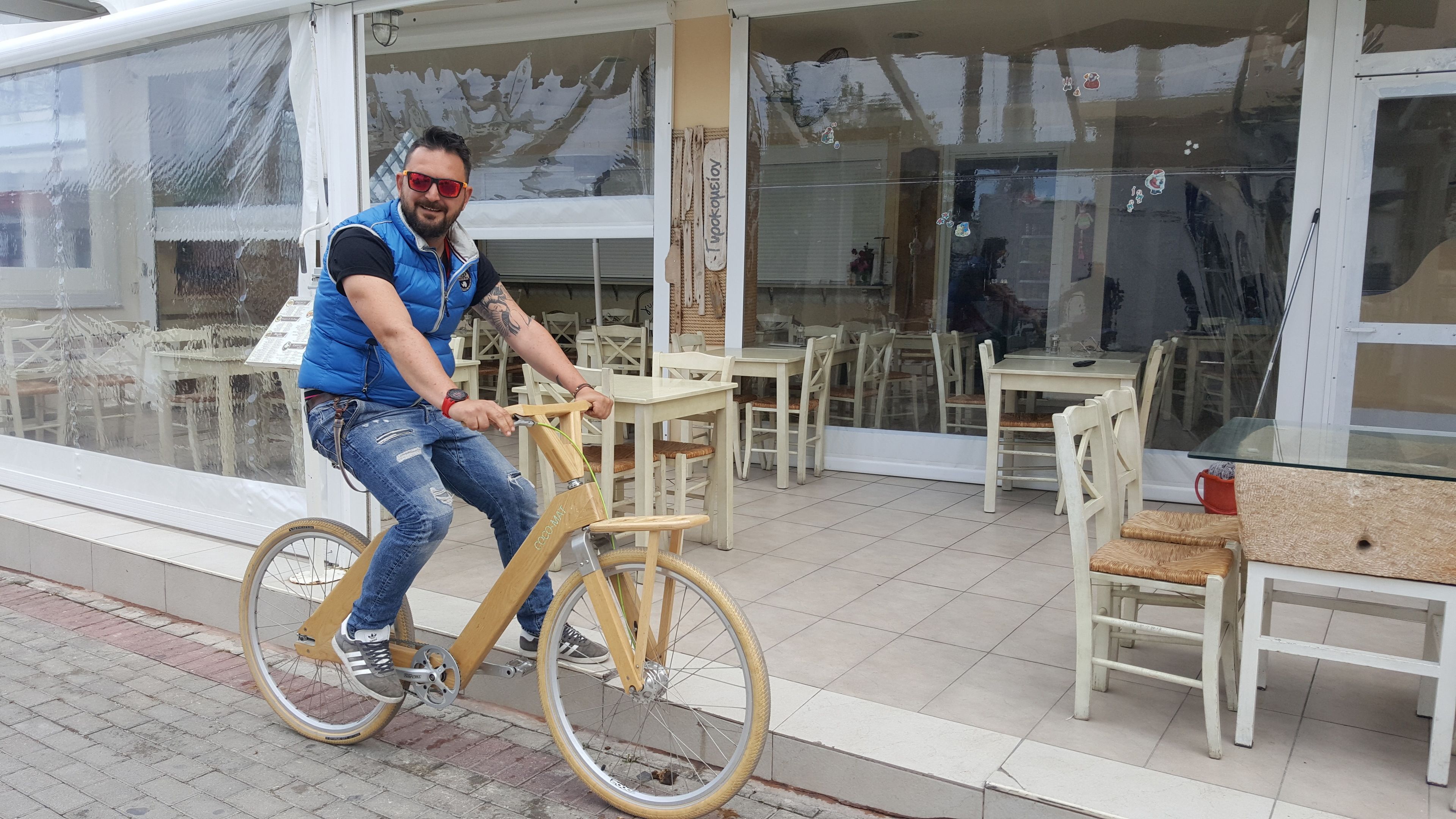 Une personne fait du vélo. Le vélo a un cadre en bois et des pédales en métal. Elle se trouve à l’extérieur d’un bâtiment doté de grandes fenêtres et d’une terrasse.