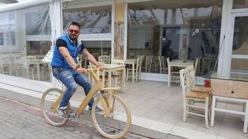 A person riding a bicycle with a wooden frame and metal pedals, outside a building with large windows and outdoor seating.